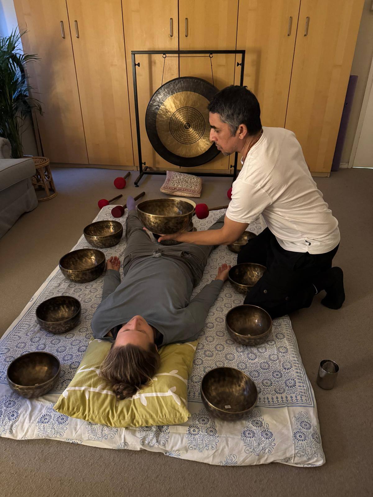 A person lies on a mat surrounded by singing bowls, while another person is seated beside them, preparing to play the bowls for sound therapy.